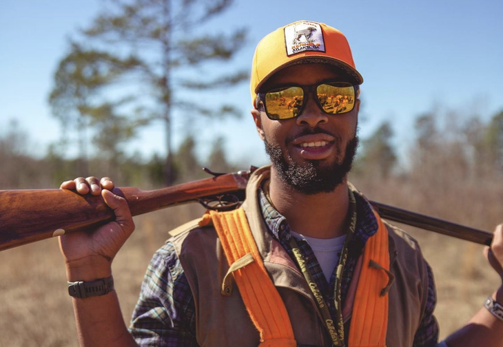 Man in sunglasses and hat holding a shotgun over his shoulders