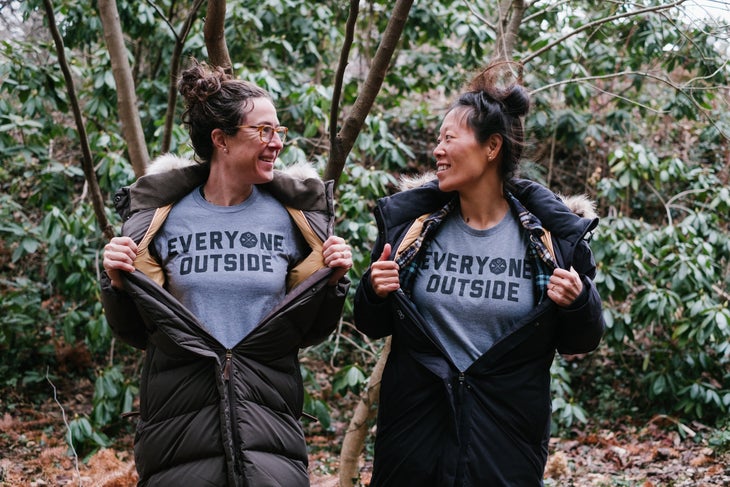 two women looking at each with jackets opened to reveal matching T-shirts that say "Everyone Outside" | Erin McGrady