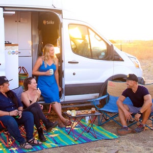 Four people relaxing in front of a white campervan in the desert, setting sun