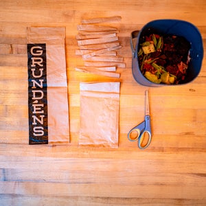 Grundens compostable packaging: A brown bag, partially cut into strips lays on a wooden surface next to a bucket of vegetable scraps and a pair of scissors