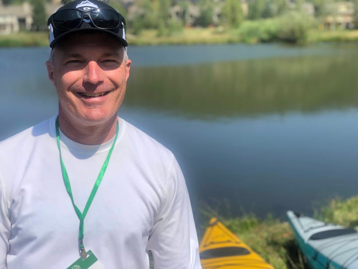 Man with green lanyard and hat smiling next to a lake