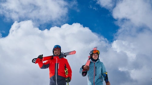 two skiers walking towards camera with skis over their shoulders