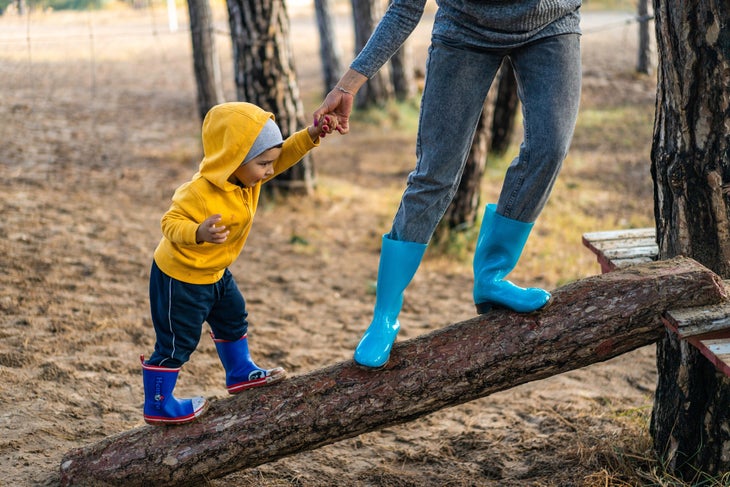 Toddler boy in yellow sweatshirt and royal blue rainboots holds the hand of an adult in gray with turquoise rain boots as they walk up a log to a…