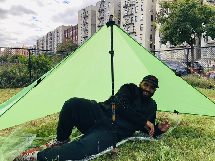 "Man wearing black lounging underneath a green tarp tent in a fenced in yard with city buildings behind him"