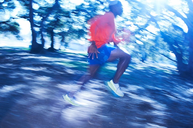 Blurred photo of Black man in blue shorts and ornge shirt running on a wooded trail