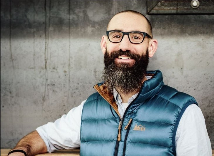 "A man with glasses, a beard, and a blue REI vest smiling at the camera."