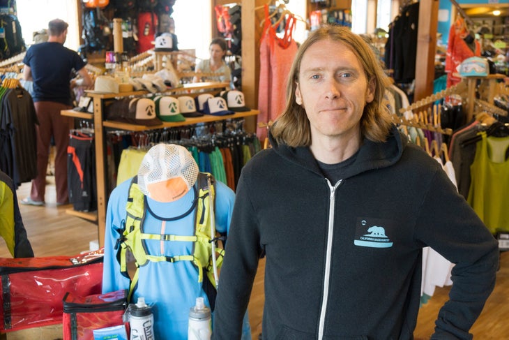 Brendan Madigan. Long haired smiling man in black hoodie standing in a retail shop