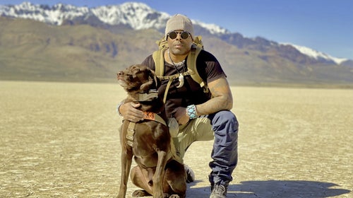 Man with backpack, sunglasses and knit beanie kneeling on a cracked desert floor with his arm around a sitting dog with snow-capped mountains in background