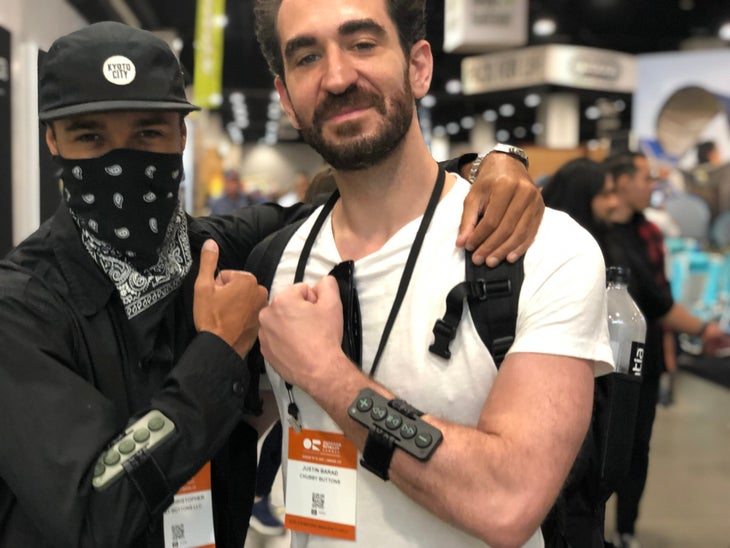 Two men, on in black shirt, cap and bandana, the other in white t-shirt bumping fists at a trade show