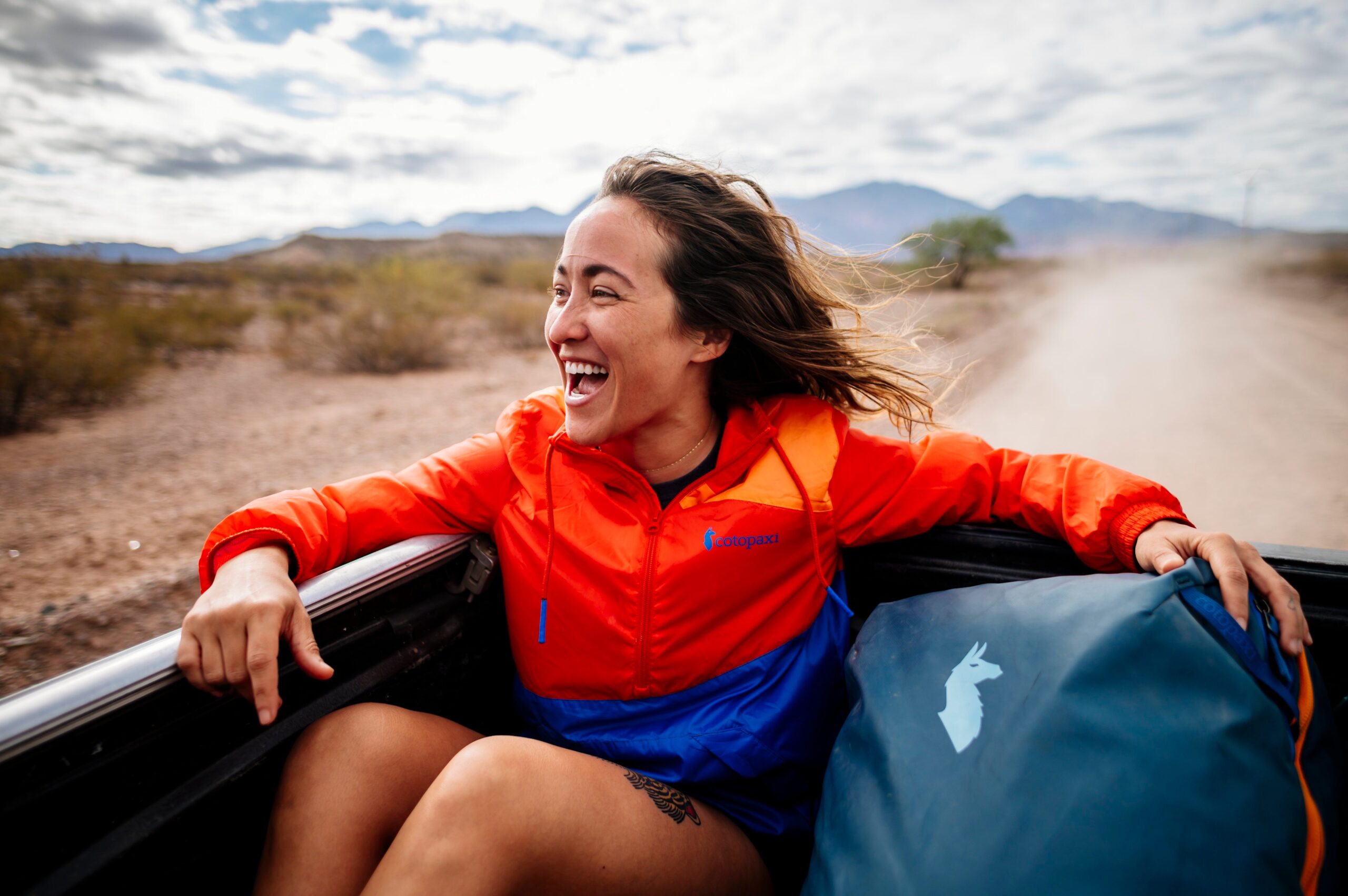 smiling woman riding in back of truck wearing orange and blue Cotopaxi jacket with desert and mountains behind her