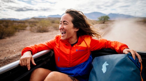 smiling woman riding in back of truck wearing orange and blue Cotopaxi jacket with desert and mountains behind her