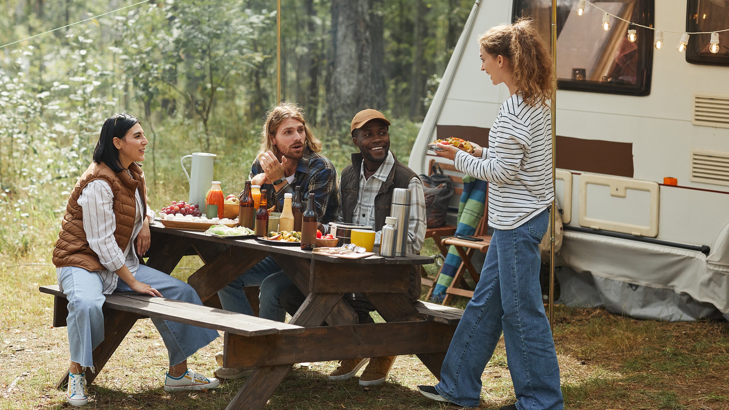 Full length view at diverse group of friends enjoying picnic outdoors at campsite with trailer van