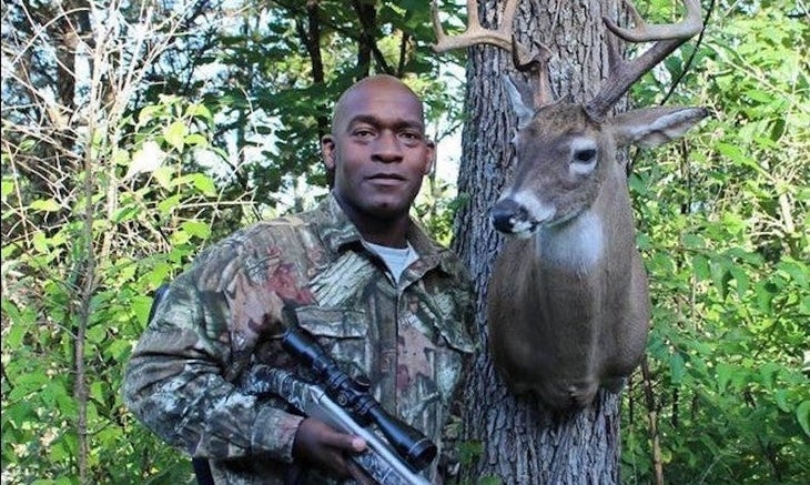 Man in hunting fatigues poses with buck head in the woods
