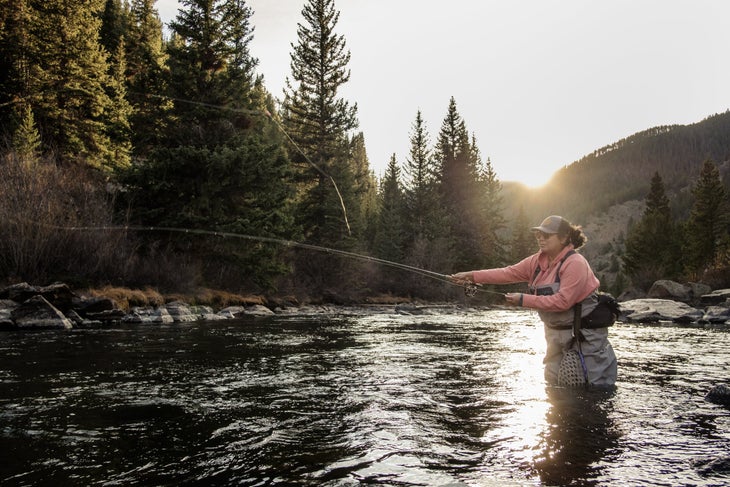 Woman in pink shirt and waders casting a fishing rod in a fiver with tall evergreen trees on the shore | hunt fish diversity