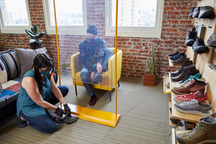 KEEN_FIT_SHIELD_0016 A retail employee helps a customer try on shoes in a store with a KEEN Fit Shield between them.