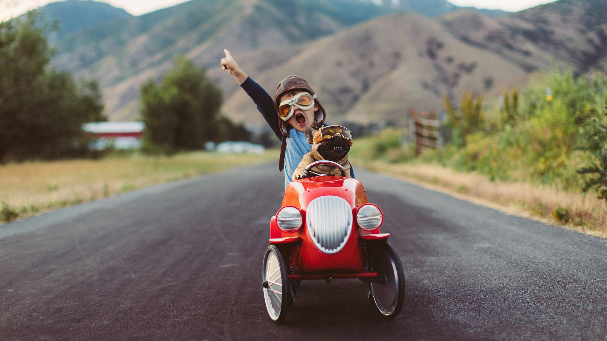 A young boy with flying goggles and flight cap races a red toy car with his pet and best friend French Bulldog along a small road in Utah.