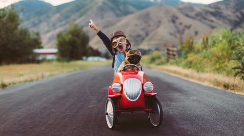 A young boy with flying goggles and flight cap races a red toy car with his pet and best friend French Bulldog along a small road in Utah.
