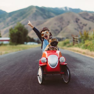 A young boy with flying goggles and flight cap races a red toy car with his pet and best friend French Bulldog along a small road in Utah.