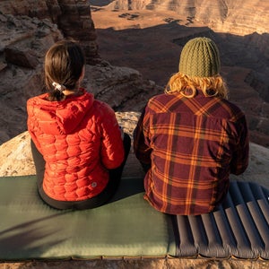 man and woman sitting side by side on a Klymit sleeping pad looking out over a canyon