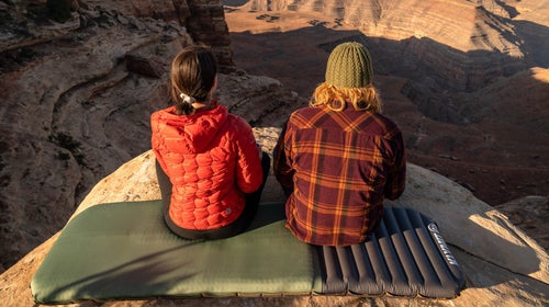 man and woman sitting side by side on a Klymit sleeping pad looking out over a canyon