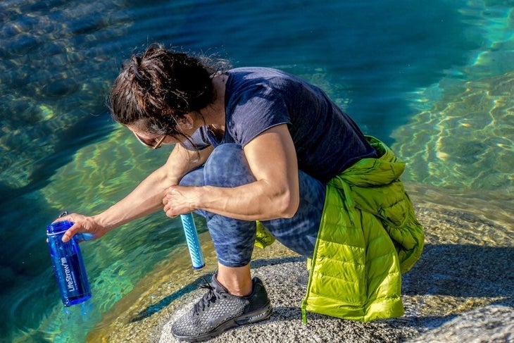 A woman kneeling to refill a LifeStraw bottle in a lake.