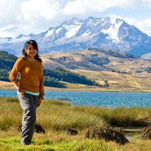 woman in gray hiking pants and orange sweater standing in the grass in front of a lake with snowy mountains in the distance