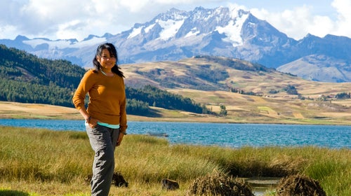 woman in gray hiking pants and orange sweater standing in the grass in front of a lake with snowy mountains in the distance
