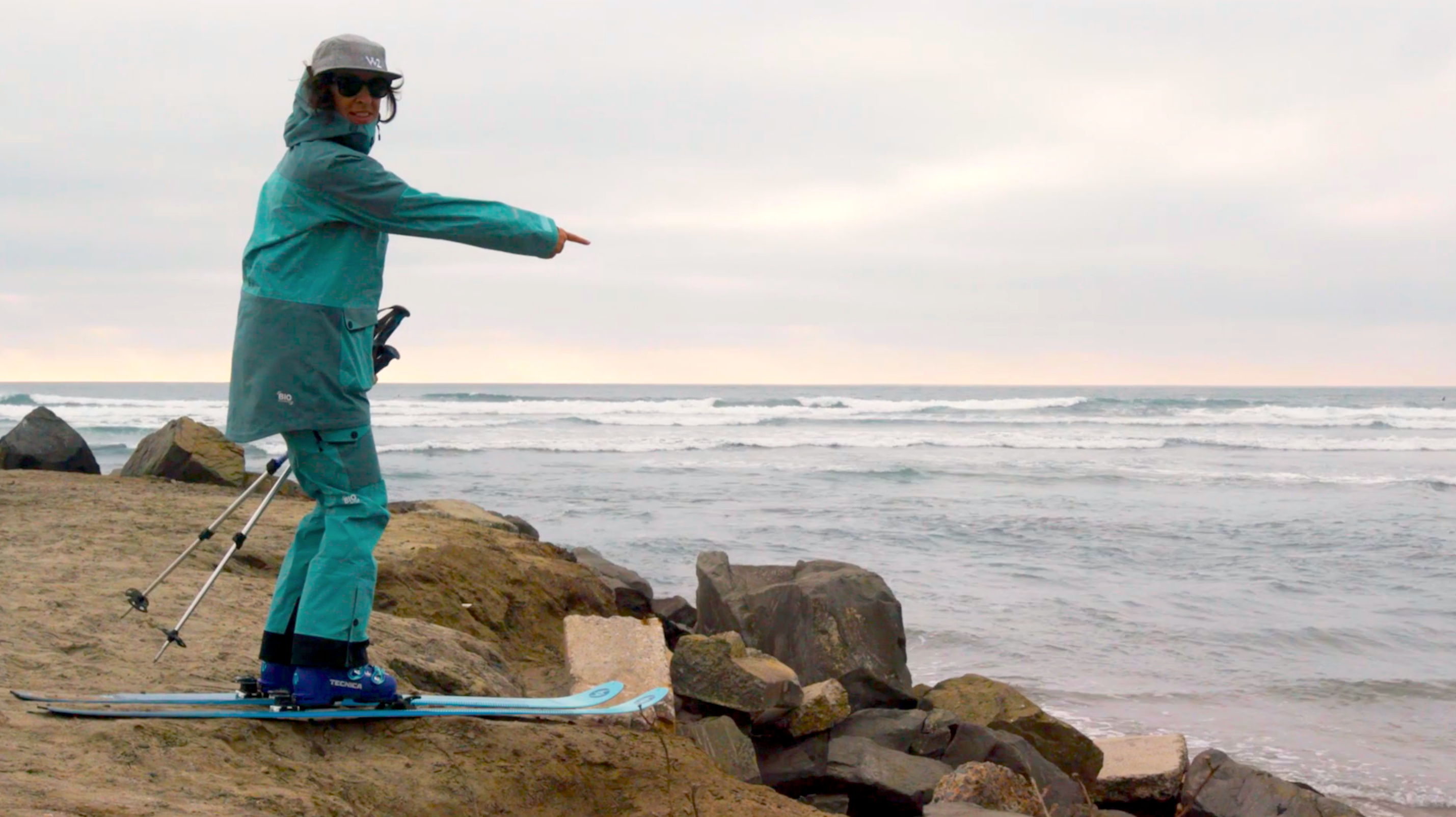 Woman wearing skis on a rocky beach pointing at the sea