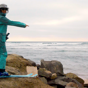 Woman wearing skis on a rocky beach pointing at the sea