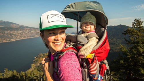 A young woman stands atop a mountain with a baby in a backpack