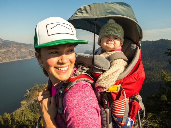 A young woman stands atop a mountain with a baby in a backpack