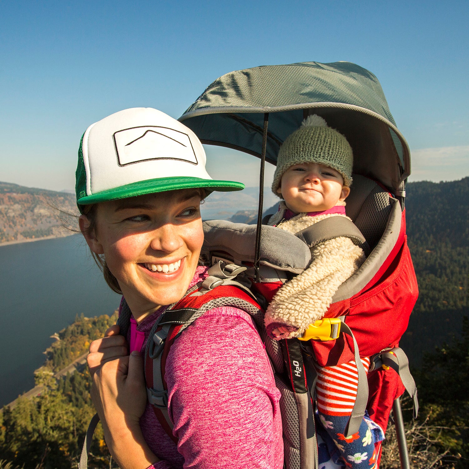 A young woman stands atop a mountain with a baby in a backpack