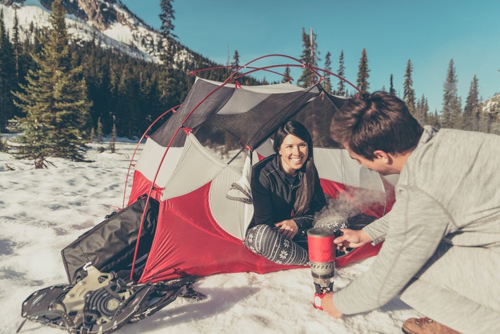 NRL_3575_Edit_2 A man boils water as a woman sits in a white and red tent. Snow shoes are to the left. Pine trees and blue sky fill the background.