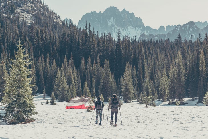 Two people snow shoe toward a forest of pine trees. A red and white tent sits in a field of snow.