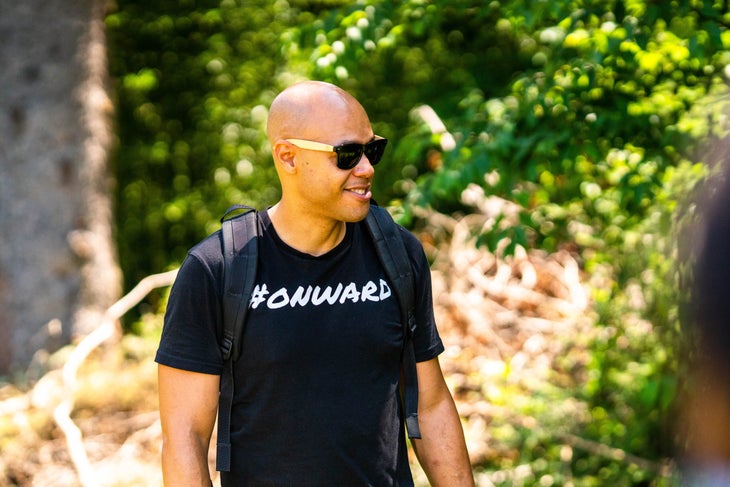 Man in black T-shirt with white letters #ONWARD and sunglasses with trees in background