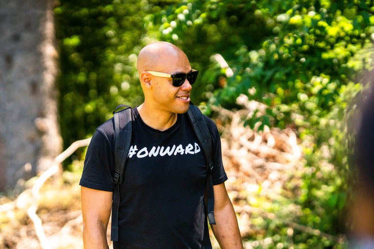 Outerthere_Al Berrios Man in black T-shirt with white letters #ONWARD and sunglasses with trees in background