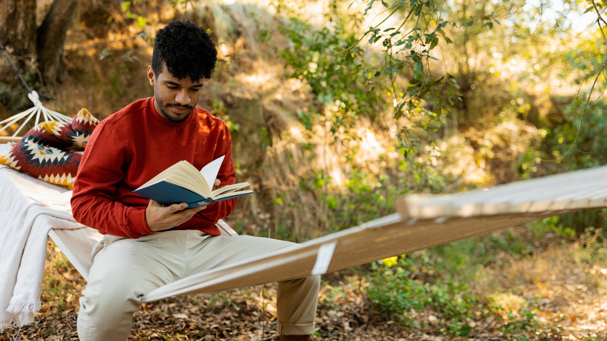 Young man sitting in a hammock reading a book while enjoying nature
