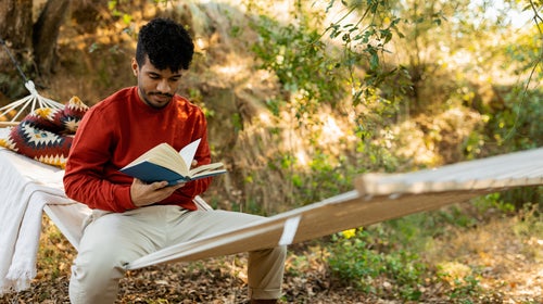 Young man sitting in a hammock reading a book while enjoying nature