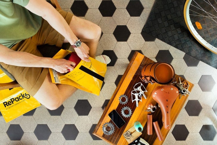 Overhead show of a person in shorts and a T-shirt putting something into a yellow mailer next to a table covered in bike parts