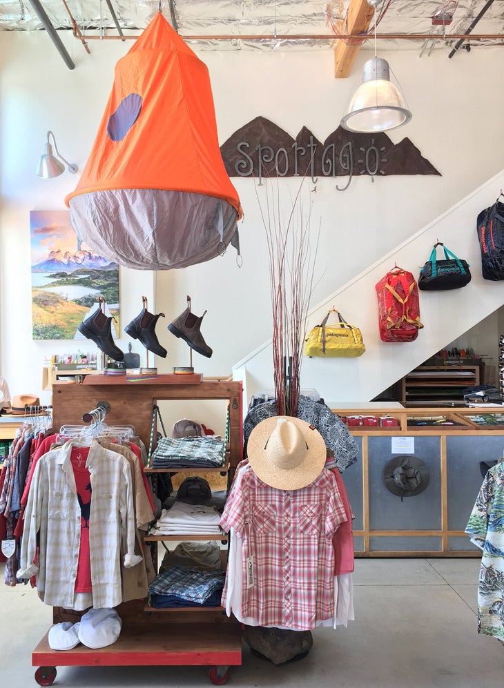 sportago Interior shot of retail store, Sportago sign on the wall, tent hanging from ceiling, racks with clothing, boots, and luggage hanging on wall.