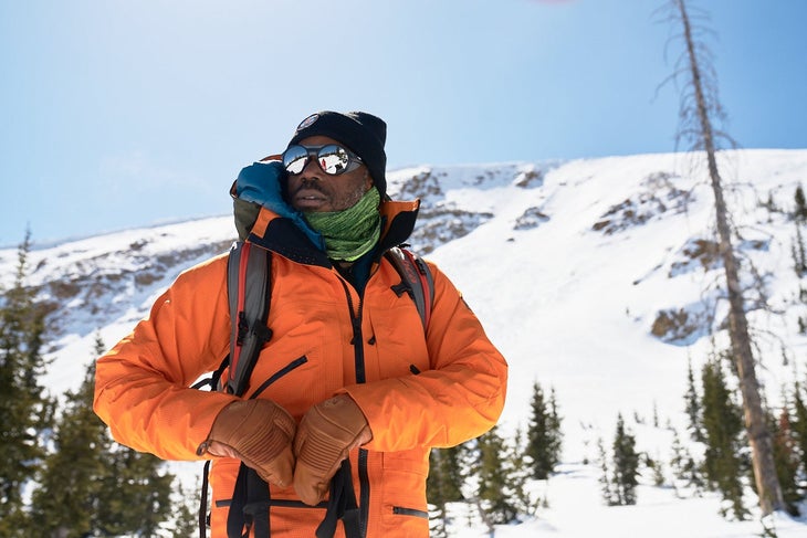 Black male skier in orange jacket, backpack, neck gaiter, mirrored sunglasses and black cap with a ski hill behind him