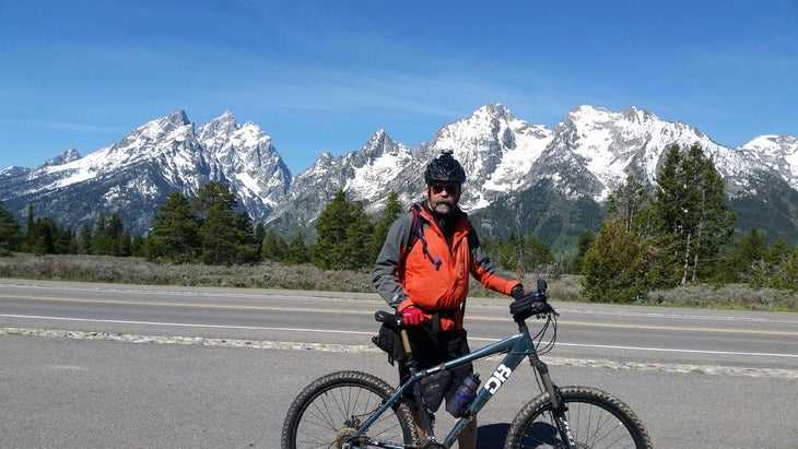 Steve Wilkinson / Man with beard and red and gray jacket standing with his green oiuntain bike on a roadside with blue sky and snow-covered mountains…