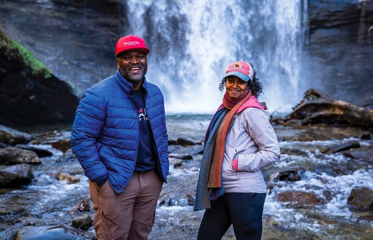 Man and a woman in hats and jackets stand in front of a waterfall