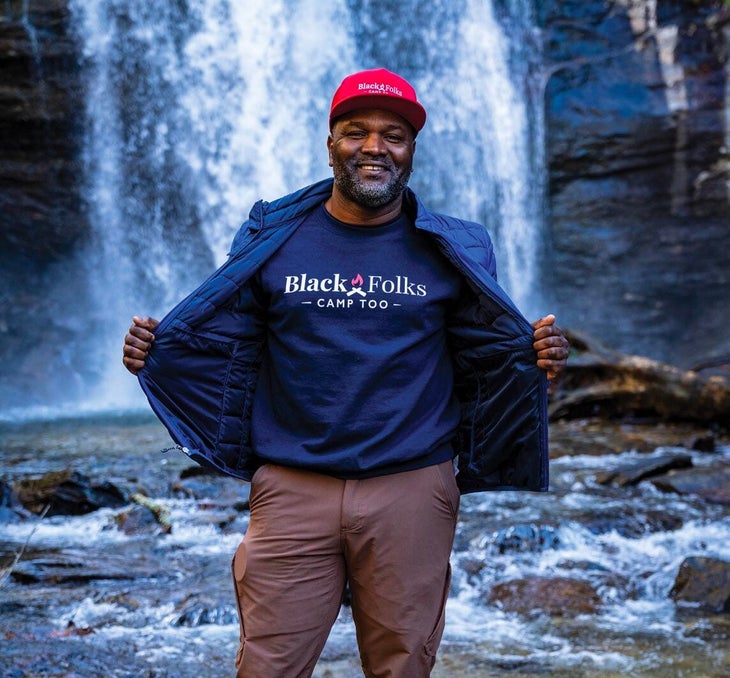"Man standing in front of a waterfall opens a jacket to reveal a T-shirt that says \"Black Folks Camp Too\""