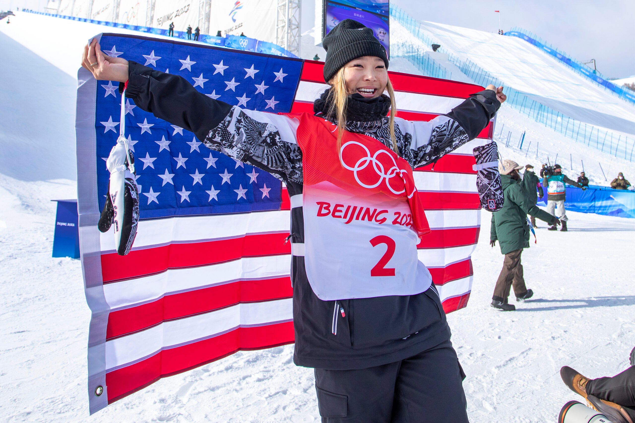 Chloe Kim smiles and holds a US flag in front of ski slopes in Zhangjiakou, China.