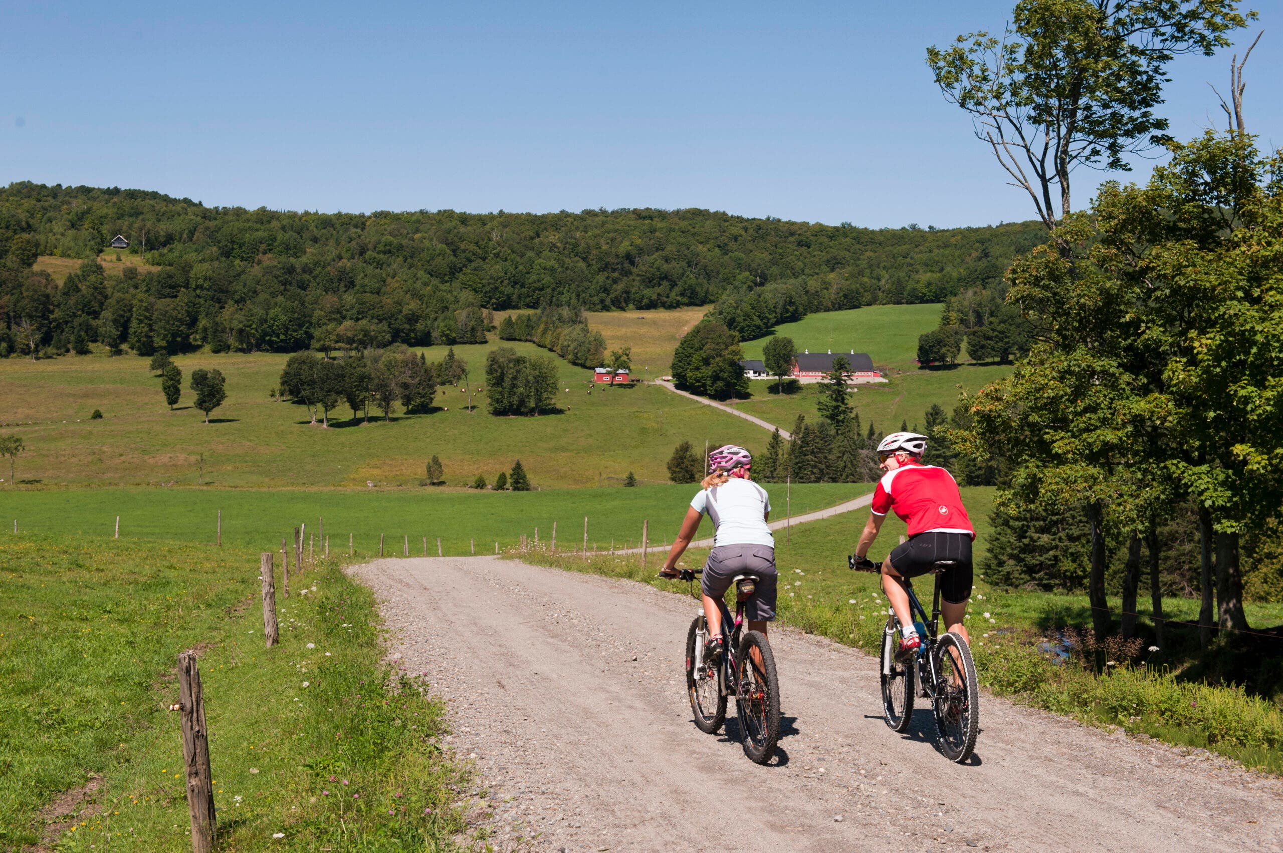 Two cyclists ride a gravel road between green fields in the Vermont country side. 