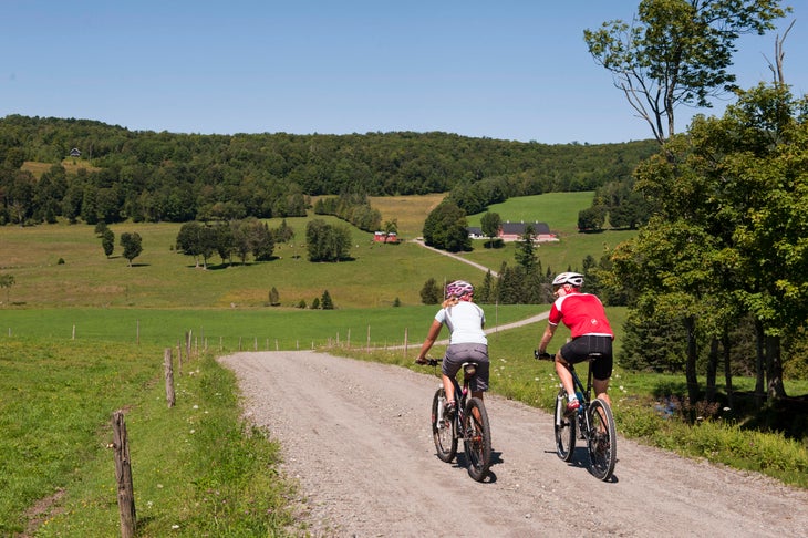 Two cyclists ride a gravel road between green fields in the Vermont country side. 