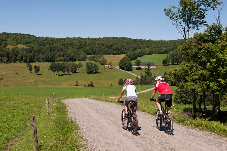 Two cyclists ride a gravel road between green fields in the Vermont country side.