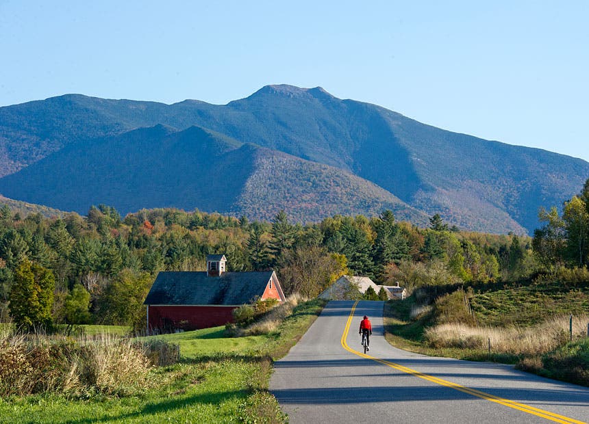 A road cyclist rides toward mountains and a red barn in Vermont.