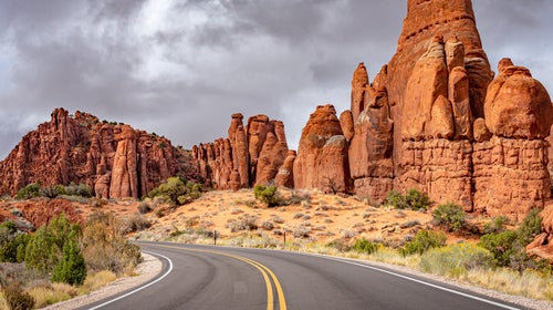 The sun brightly illuminates the red rock formations of the Fiery Furnace area of Arches National Park, contrasted by dark storm clouds in the background. The park road can be seen in the foreground.